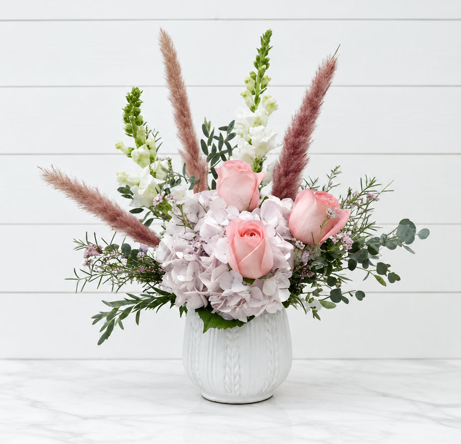 A floral arrangement in a white textured ceramic vase, featuring pink roses, light purple hydrangeas, white snapdragons, and pampas grass, set against a white wood paneled wall and marble surface for new baby girl.