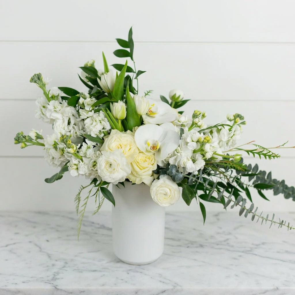 A white vase holds a lush arrangement of white roses, orchids, tulips, and greenery.