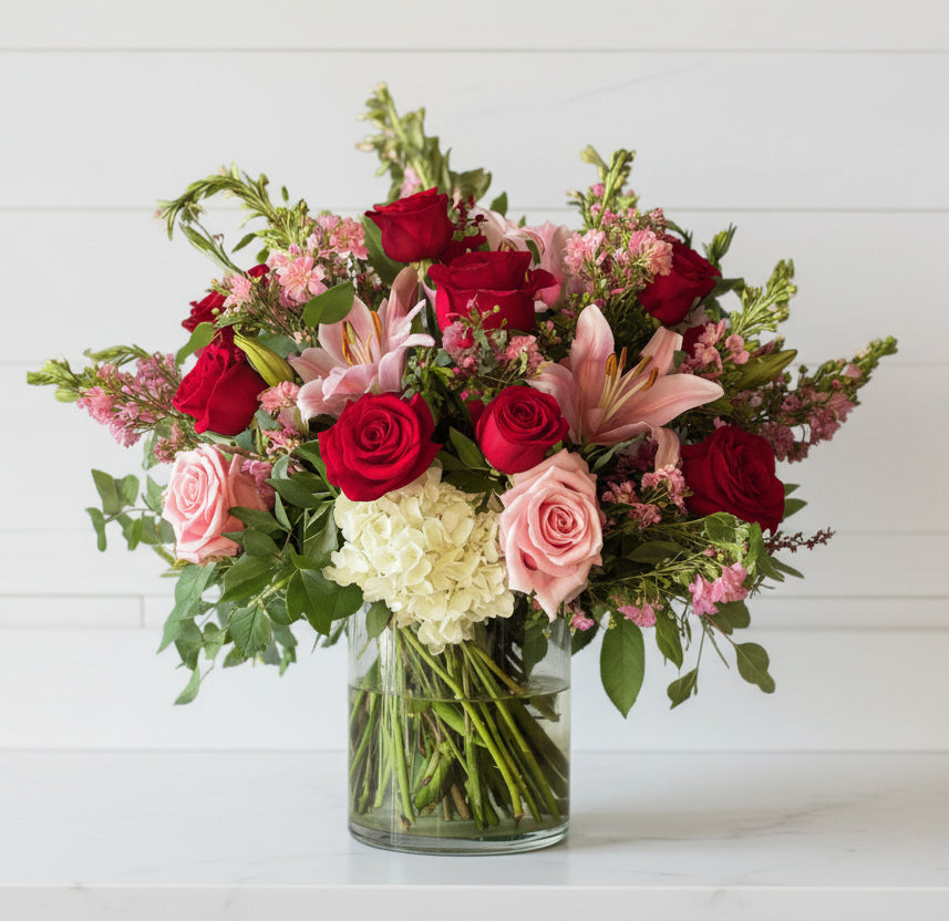 Bouquet of red roses, pink lilies, and white hydrangeas in a clear glass vase on a light gray background