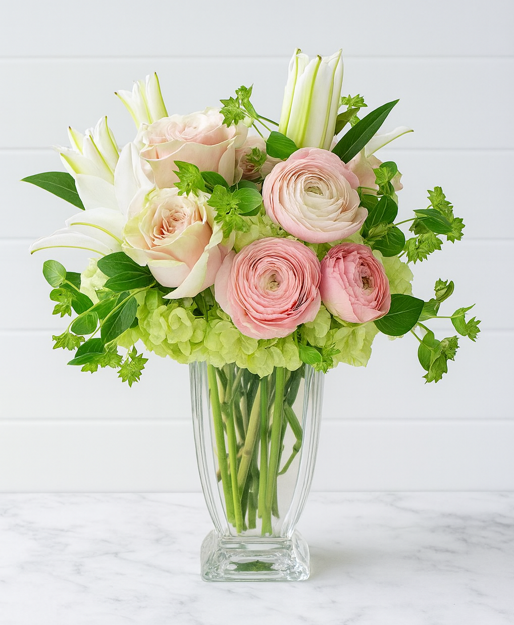 A bouquet of pink ranunculus and roses with white lilies in a glass vase.