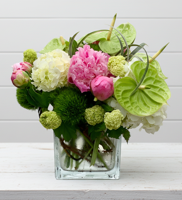 Floral arrangement with pink peonies, green hydrangeas, and white flowers in a glass vase on a light wooden background