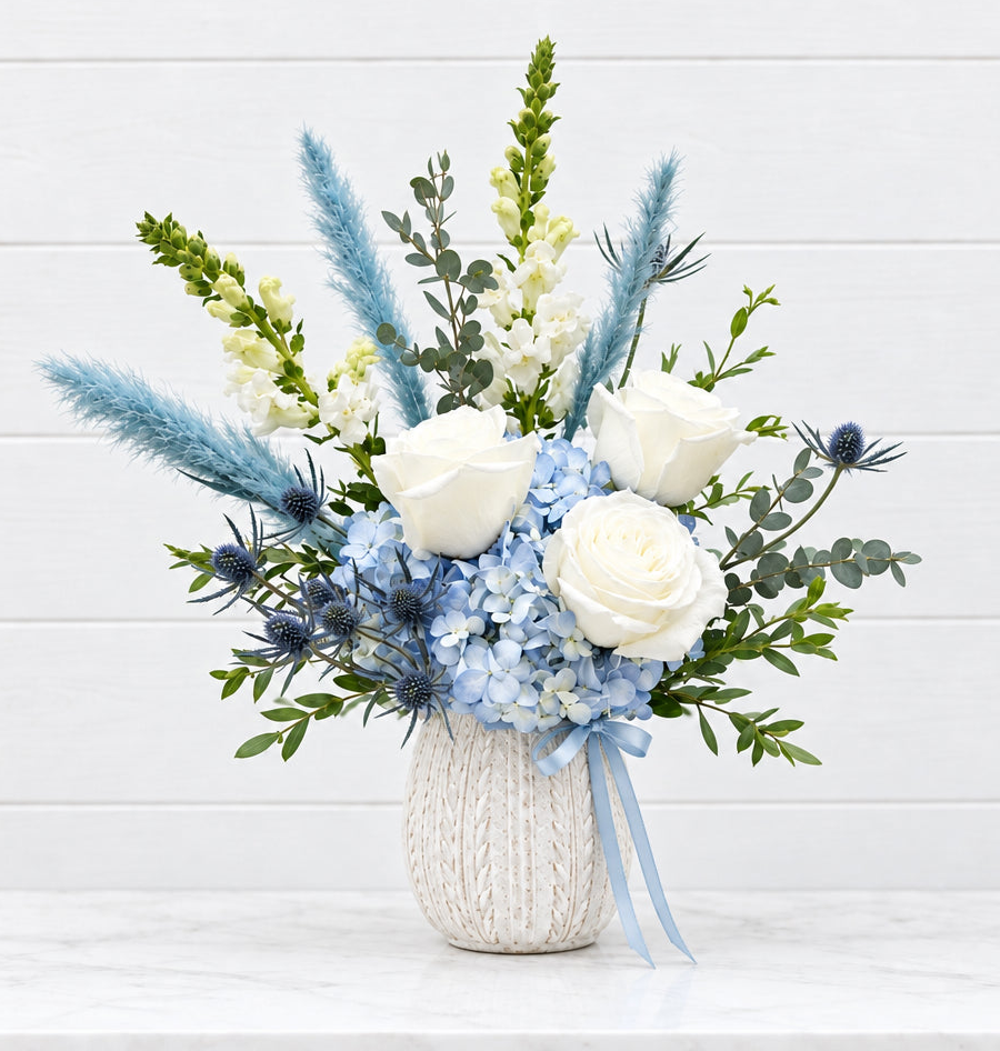 A floral arrangement in a cream-colored textured vase, featuring white roses, light blue hydrangeas, blue thistles, and blue pampas grass, set against a white paneled wall background for new baby boy welcome.