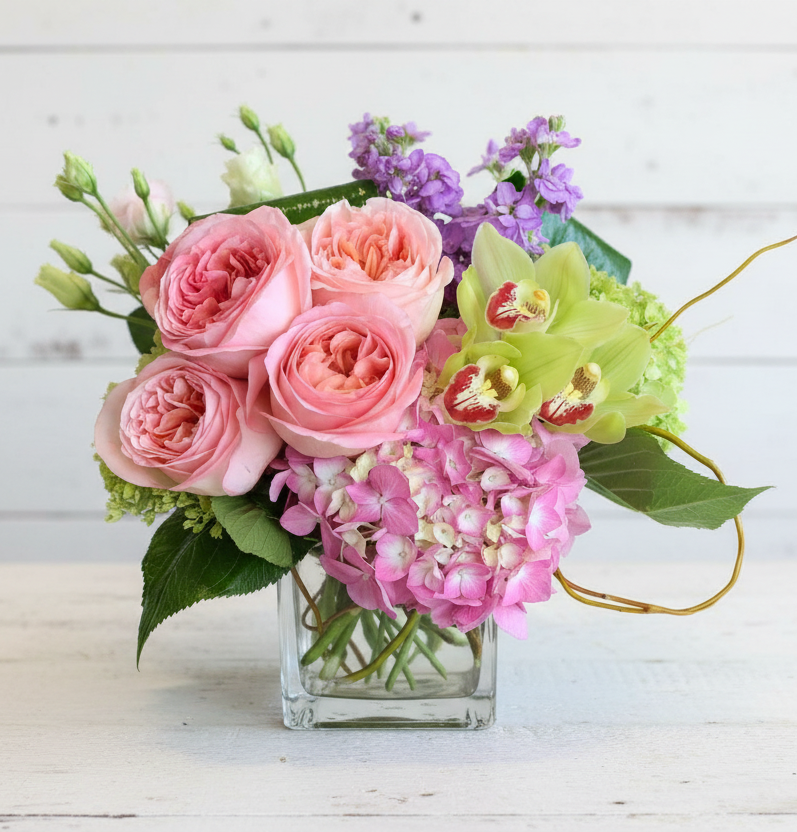 A vibrant floral arrangement featuring pink roses, green orchids, and pink hydrangeas in a glass vase.