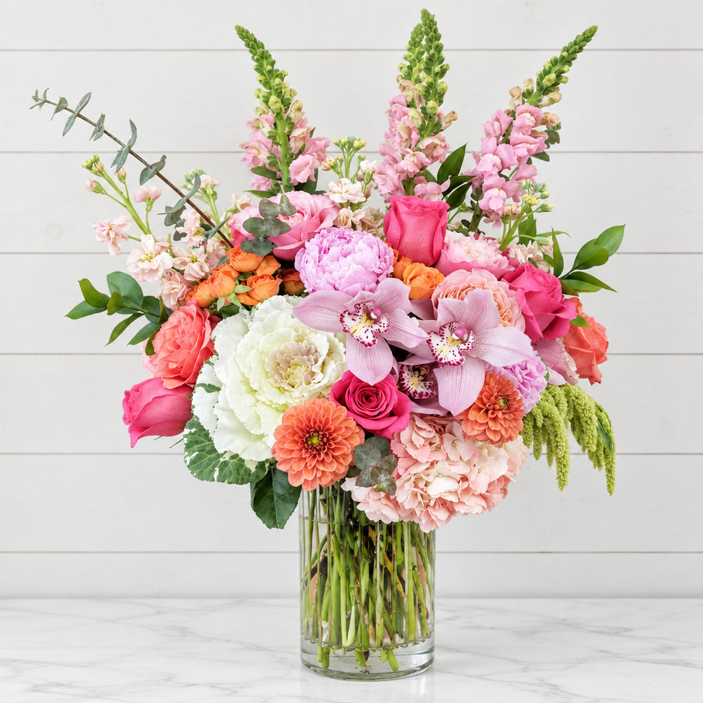 A vibrant mixed floral arrangement in a clear glass vase, featuring pink roses, light pink hydrangeas, white carnations, orange zinnias, and green foliage, displayed on a peach-colored surface against a beige wall.