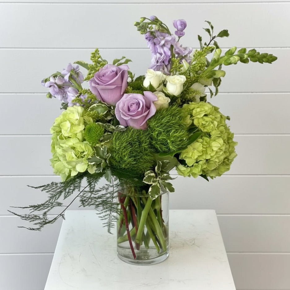 Bouquet of flowers including green hydrangeas, purple roses, and white flowers in a clear vase on a white surface with a light gray background.