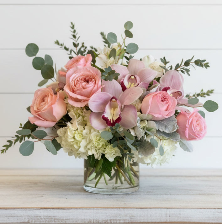 A pink floral arrangement with roses, orchids, and hydrangeas in a glass vase.