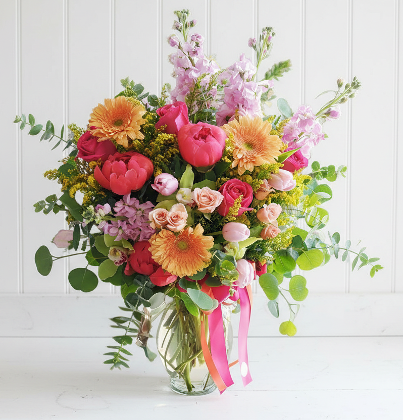 A vibrant bouquet of pink peonies, orange gerbera daisies, and yellow flowers in a glass vase.