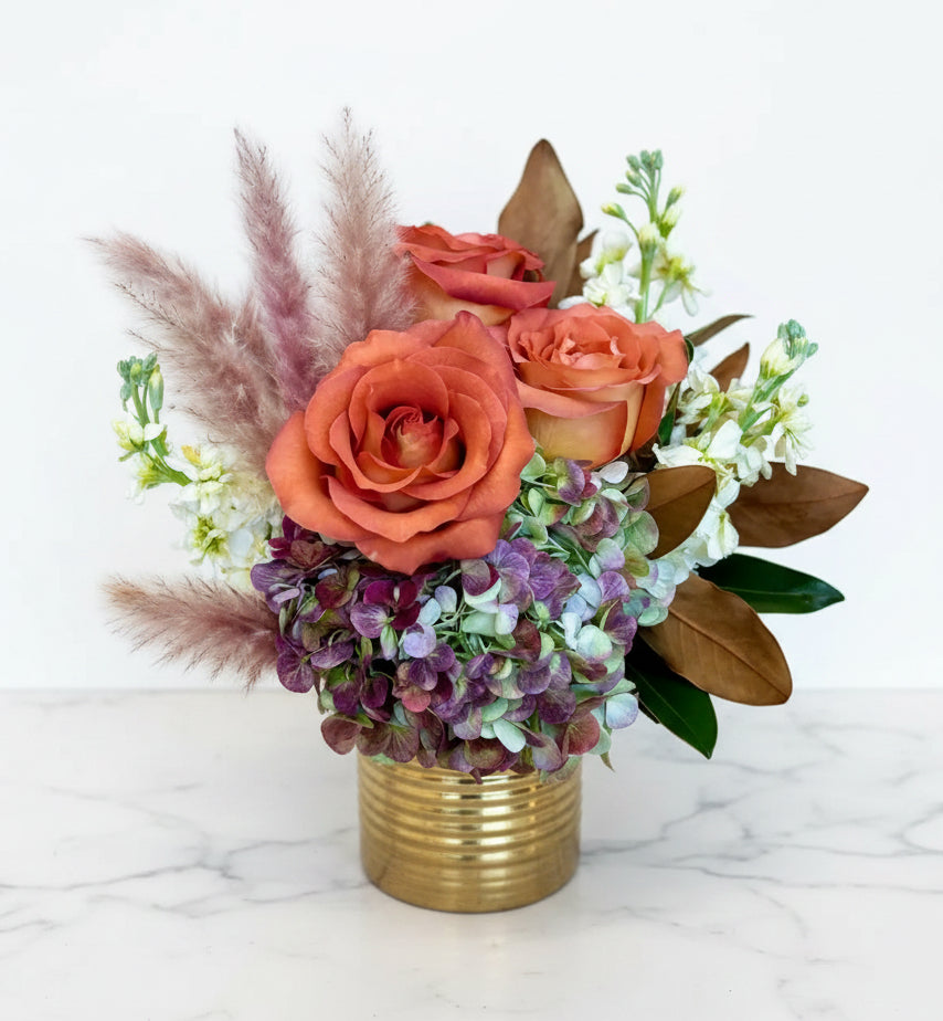Floral arrangement with coral roses, greenery, and pampas grass in a gold vase on a wooden surface.