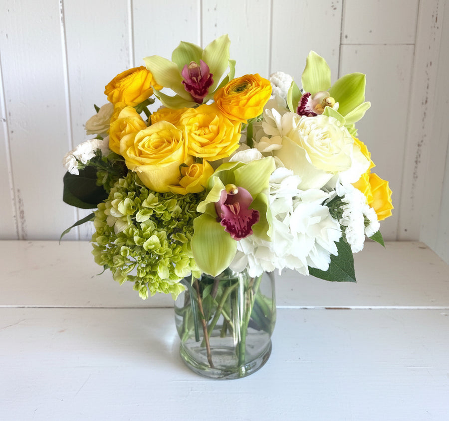 A bouquet of yellow roses, white hydrangeas, and green orchids in a glass vase.