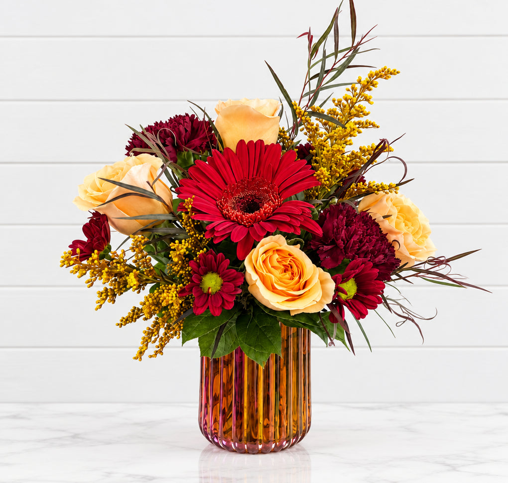 A vibrant autumn floral arrangement in a ribbed amber glass vase, featuring red and yellow roses, burgundy chrysanthemums, and yellow berries on a white pedestal against a light gray background.