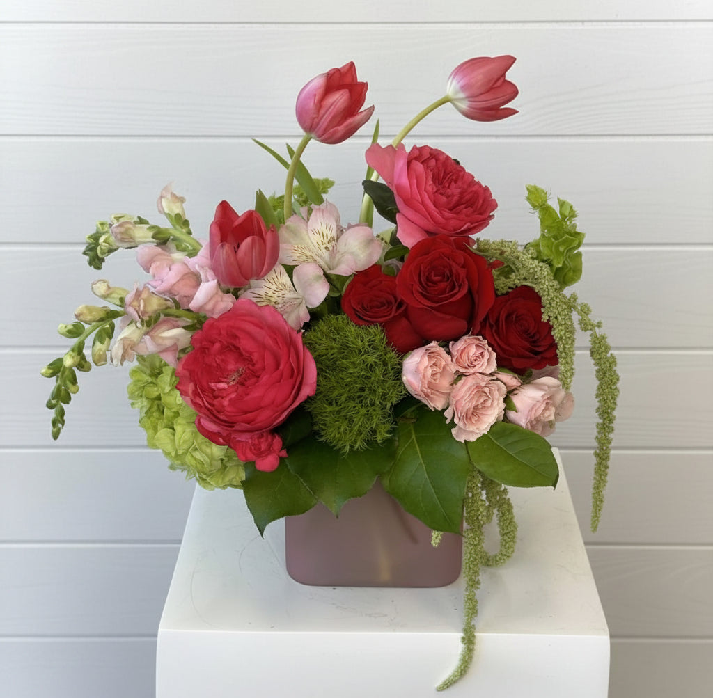 Colorful flower arrangement with red, pink, and green flowers in a vase on a white surface.
