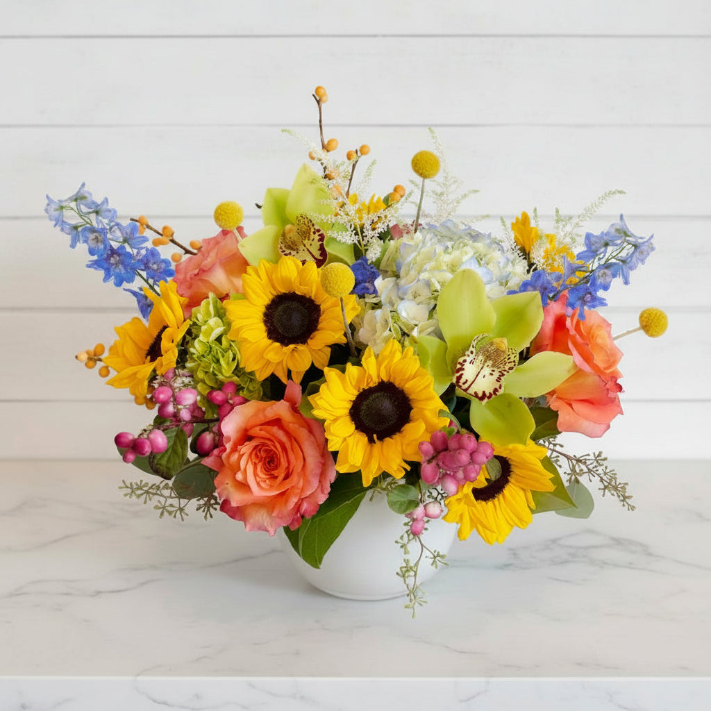Bouquet of sunflowers, roses, and other flowers in a white vase on a light wooden surface.