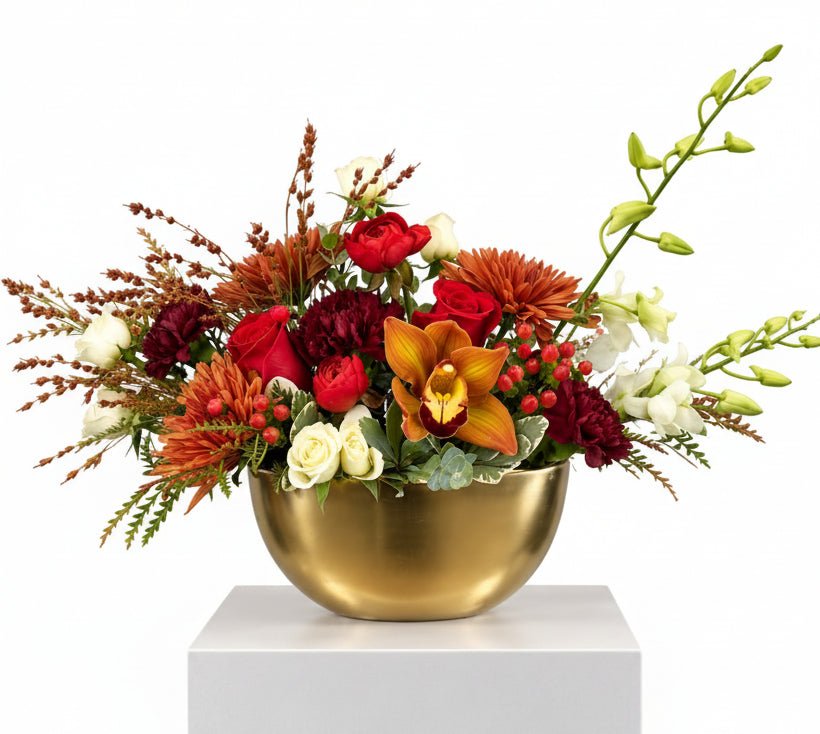 A floral arrangement in a metallic gold bowl vase, displayed on a white pedestal against a light gray studio background. The bouquet features red and burgundy flowers, orange chrysanthemums, white roses, green foliage, and a prominent orange orchid.