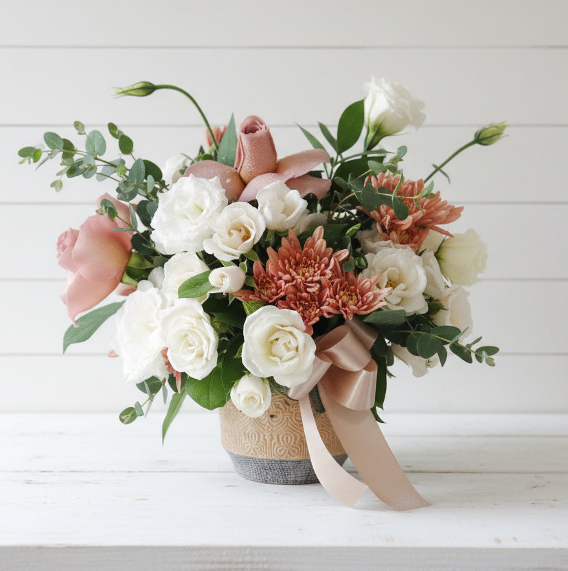 A floral arrangement with pink roses, white roses, and coral chrysanthemums in a textured pot with a ribbon.