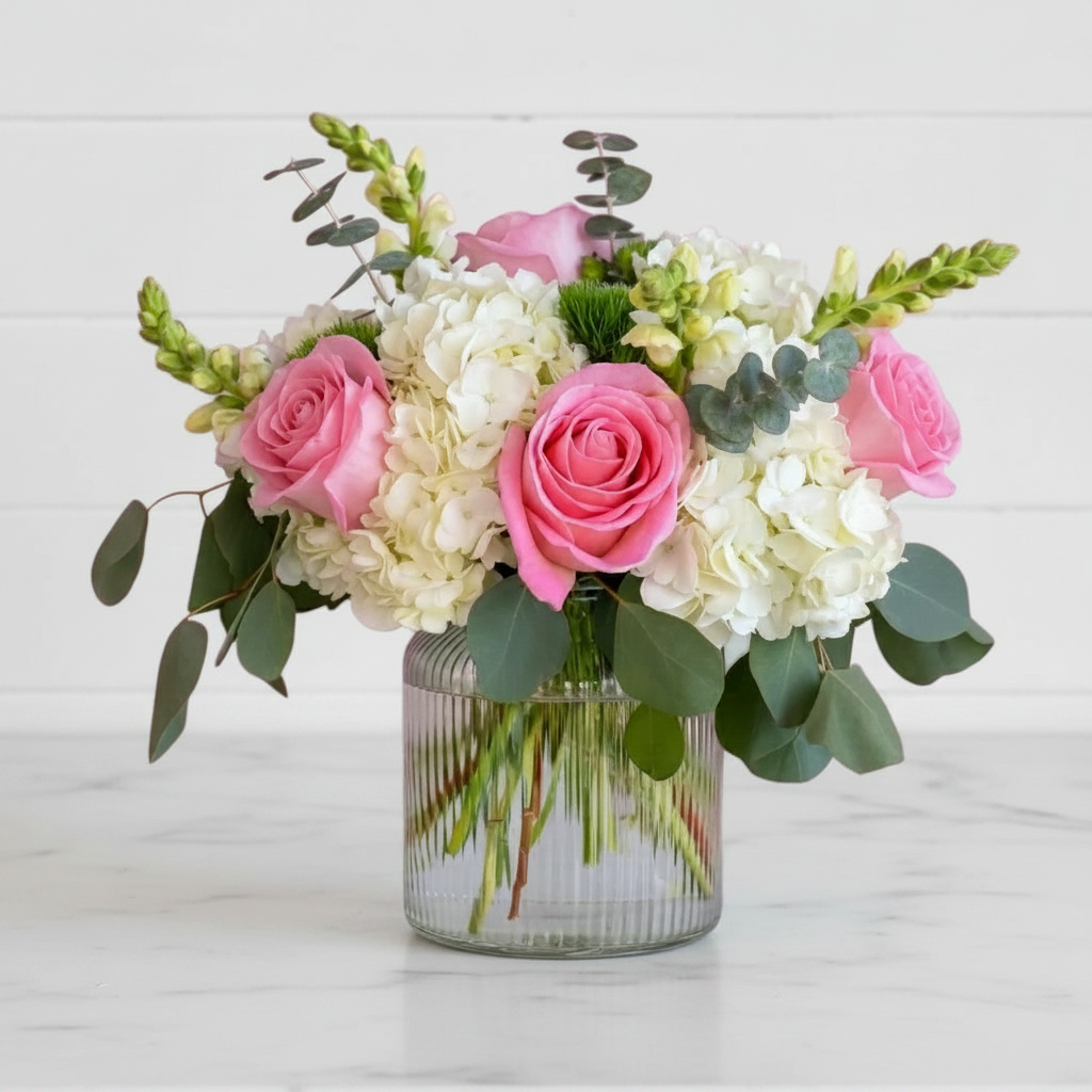 Bouquet of pink and white flowers in a clear vase on a light wooden surface.