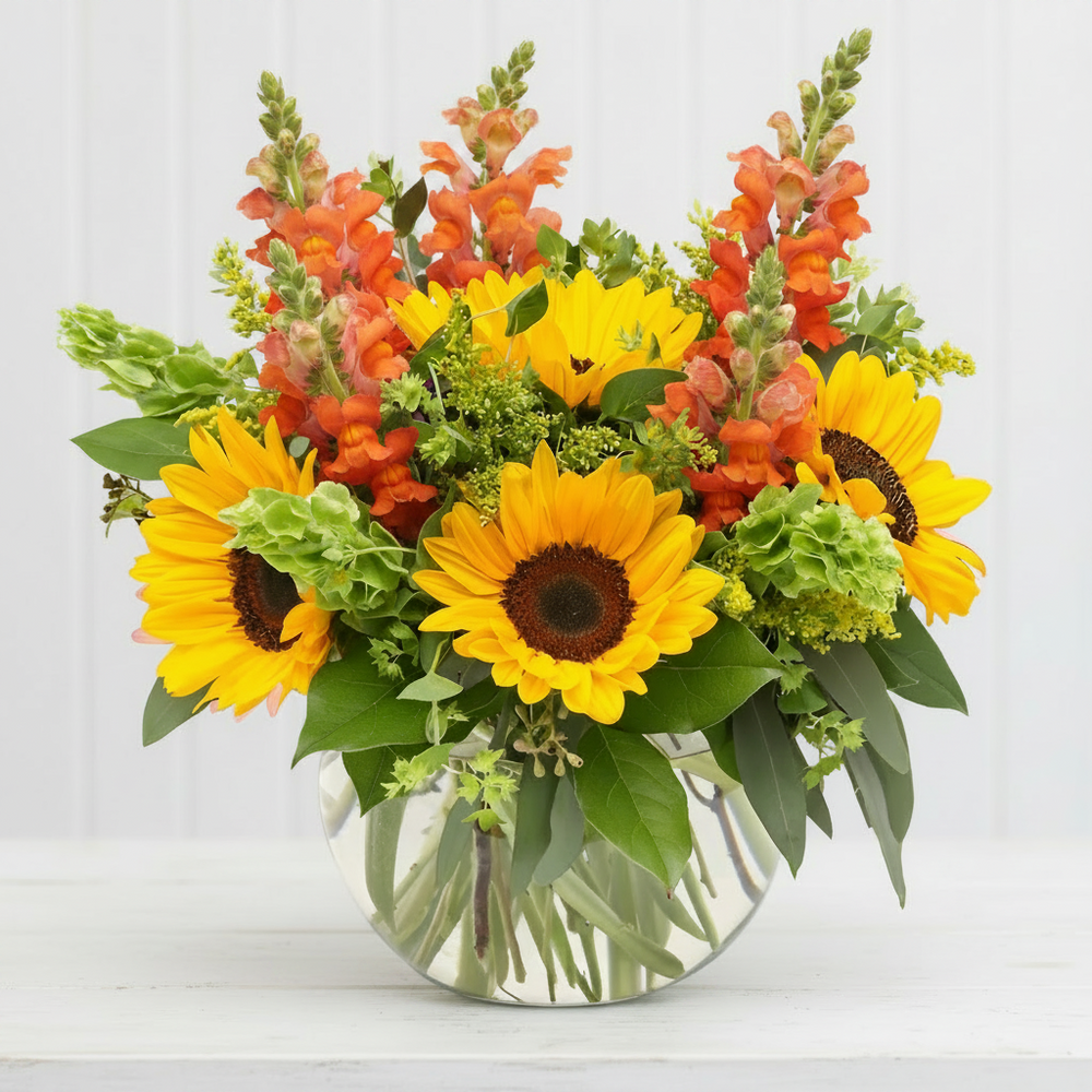 A vibrant floral arrangement featuring yellow sunflowers, orange snapdragons, and green foliage in a round clear glass vase, set against a clean white background.
