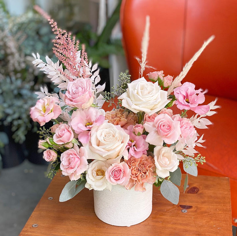 A white textured vase holds a mixed floral bouquet of pink and white roses, carnations, and other delicate blooms.