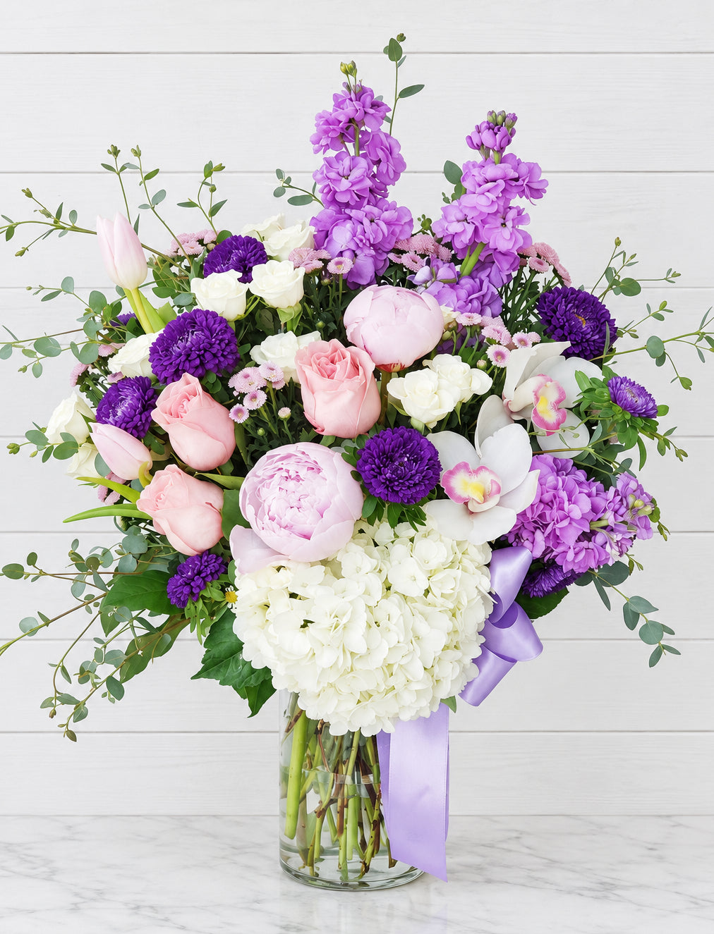 A lush bouquet of pink and white roses, purple asters, white hydrangeas, and purple stock flowers in a glass vase.